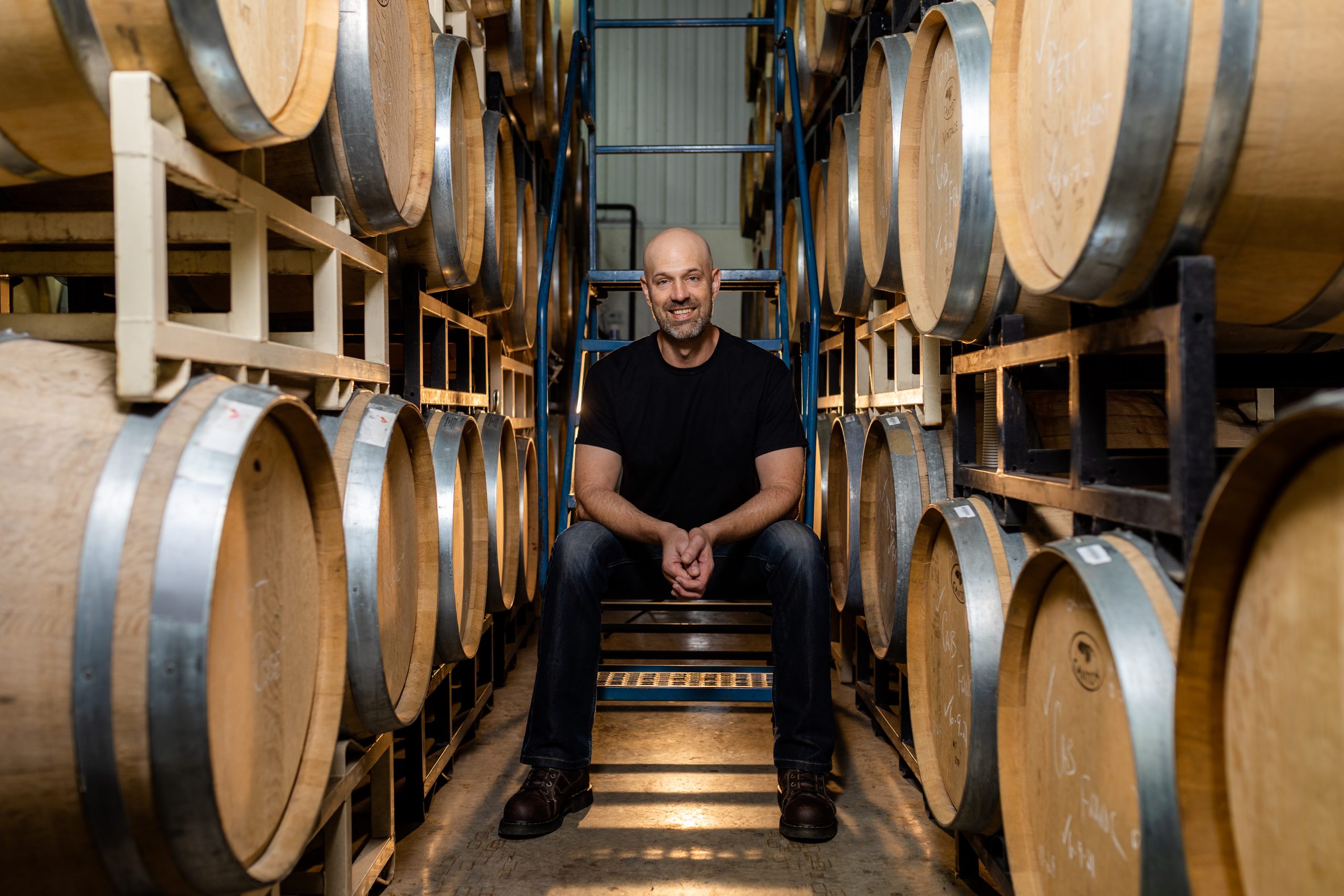 A man sitting between rows of wine barrels.